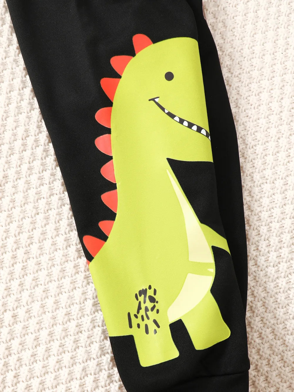Green dinosaur graphic with red spikes on black fabric against a textured beige background.