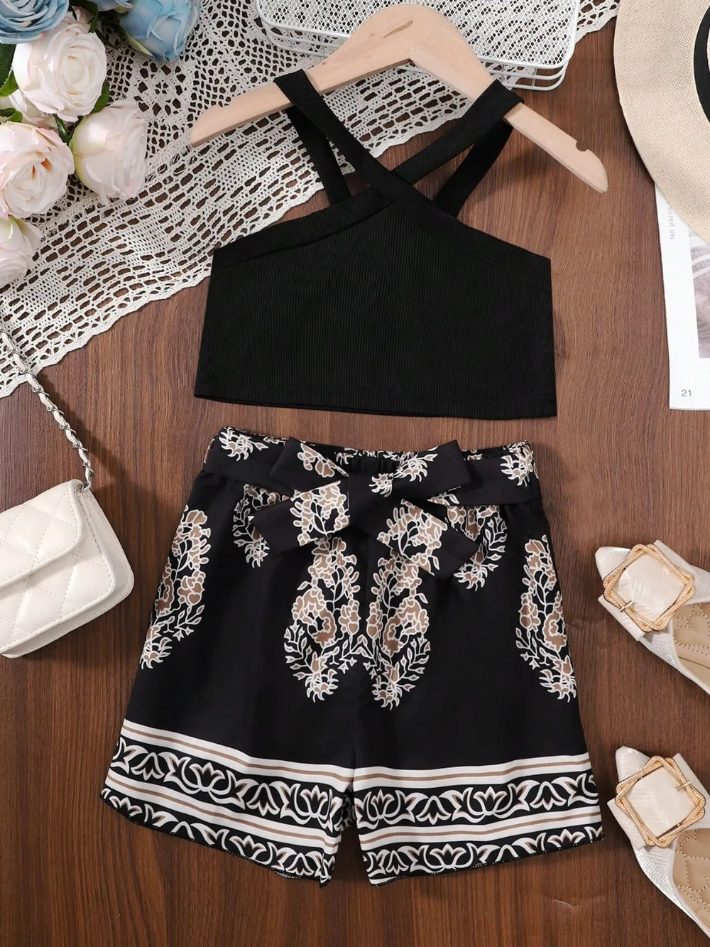Black crisscross crop top with floral patterned shorts, white handbag, and sandals on a wooden surface.