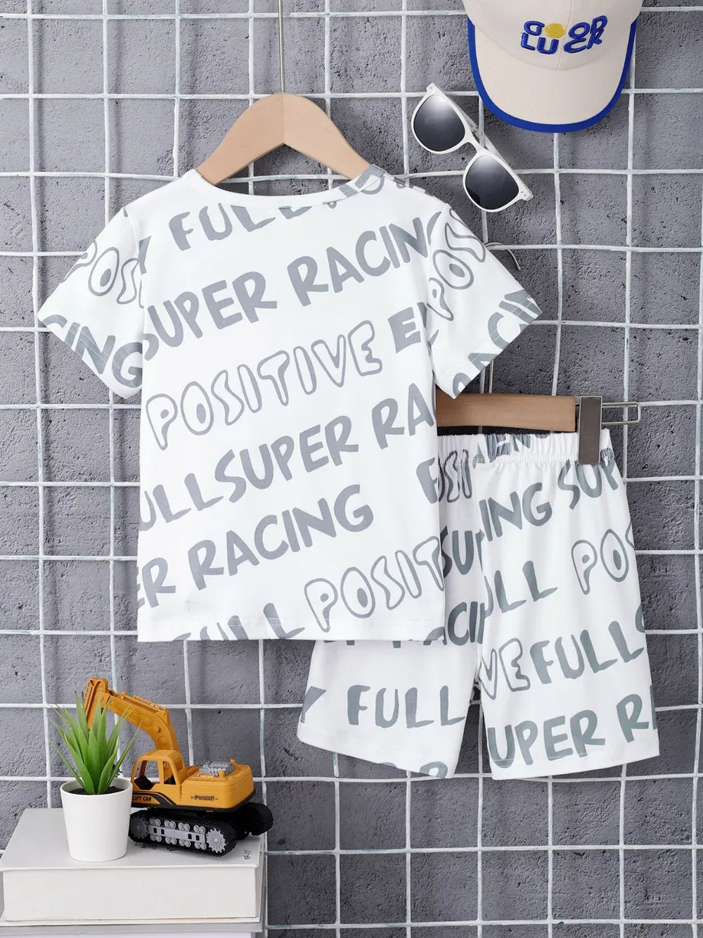 White children's t-shirt and shorts with print, displayed alongside sunglasses and a cap.