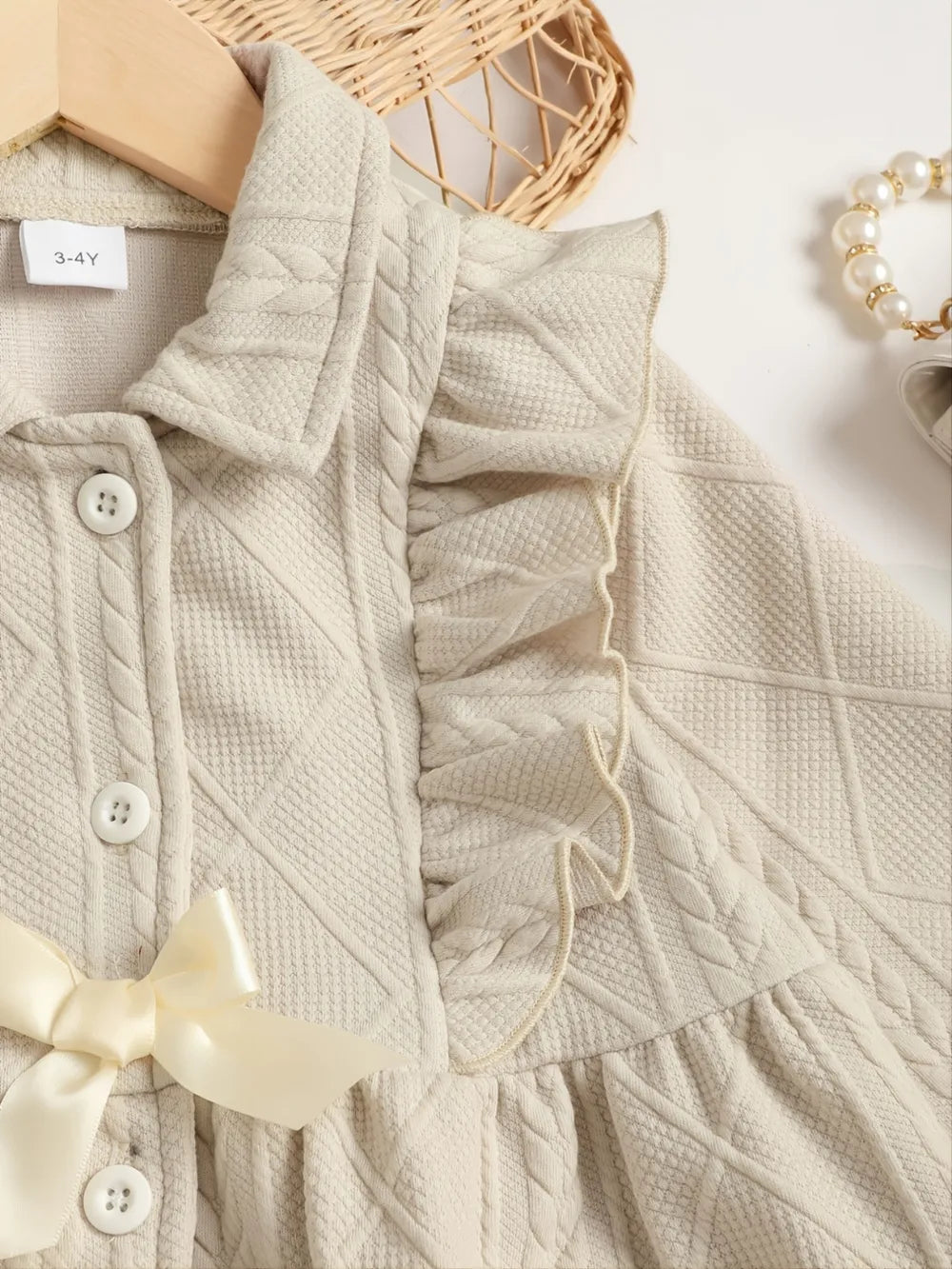 Beige infant dress with ruffles, buttons, and a bow, surrounded by accessories and decor items.