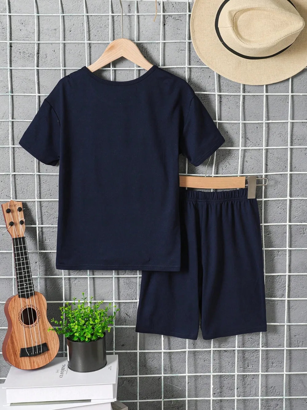 Navy t-shirt and shorts hanging on a grid, with a hat, ukulele, and potted plant nearby.