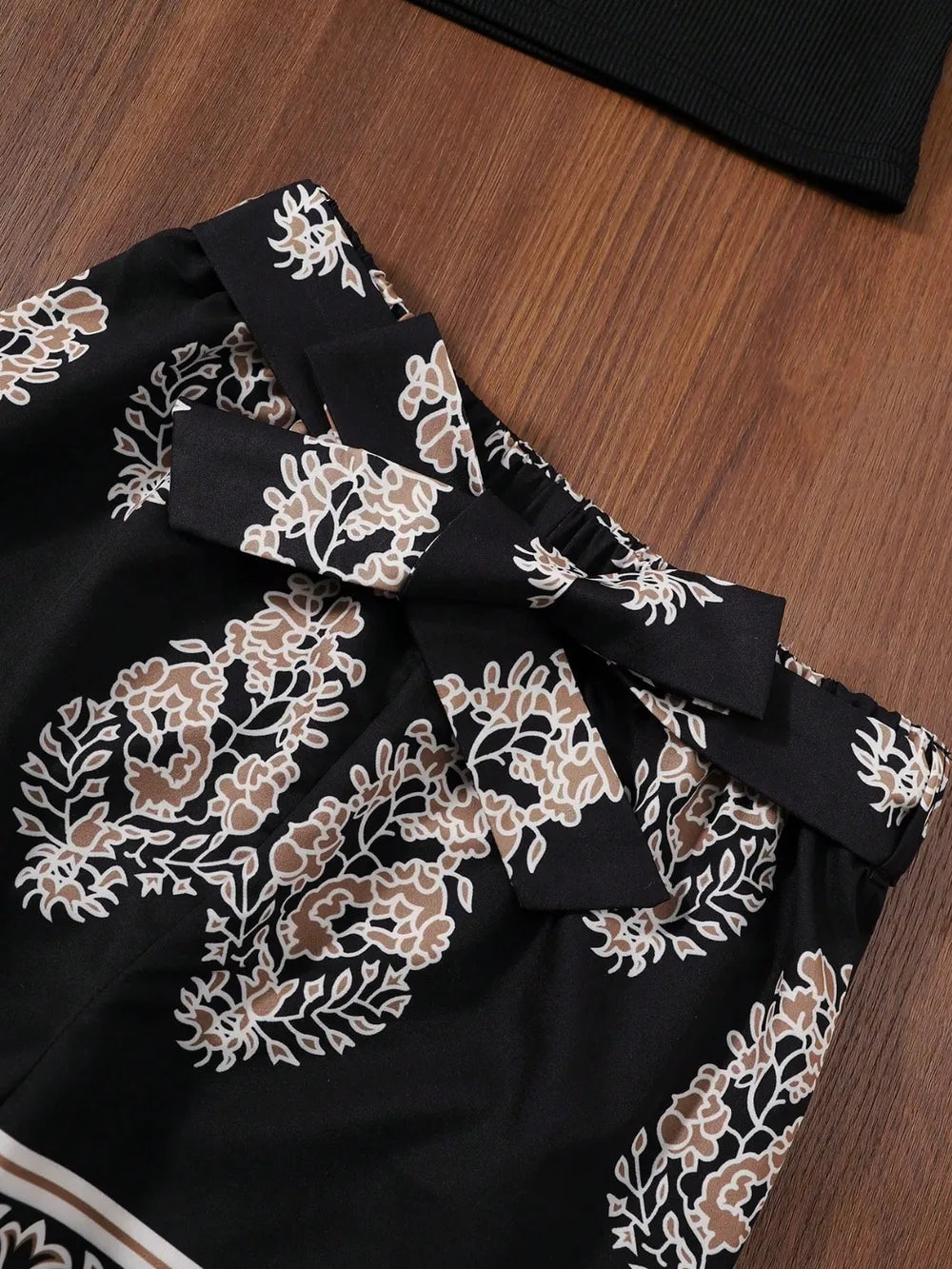 Black floral shorts with beige and white designs, featuring a tied waistband on a wooden surface.