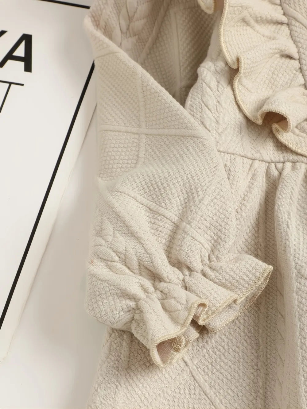 Light beige children's jacket with ruffled sleeves and decorative buttons on a wooden hanger.