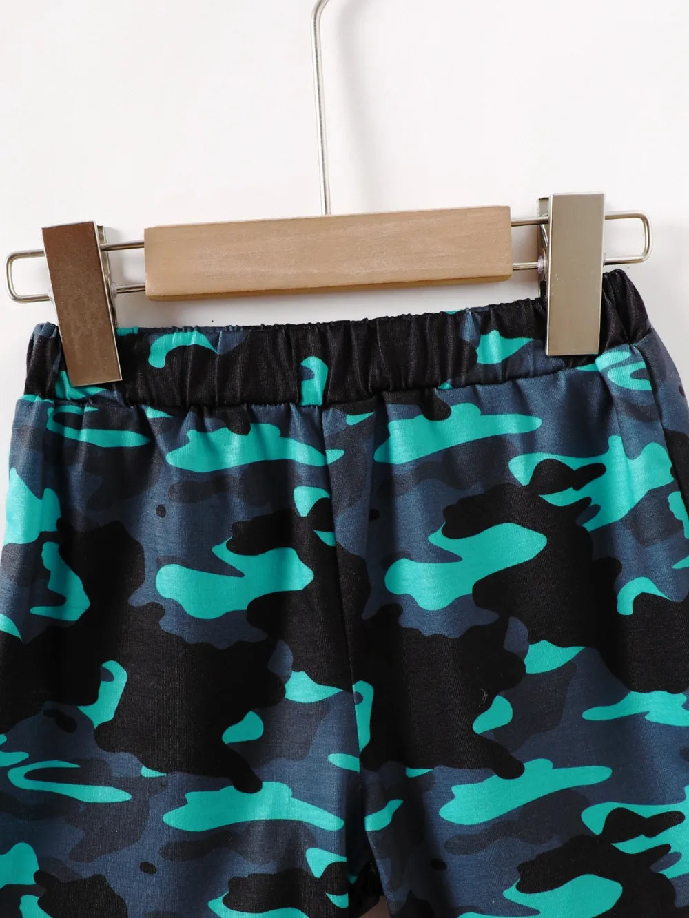 Camo-patterned shorts in shades of teal and black hanging on a wooden clip.
