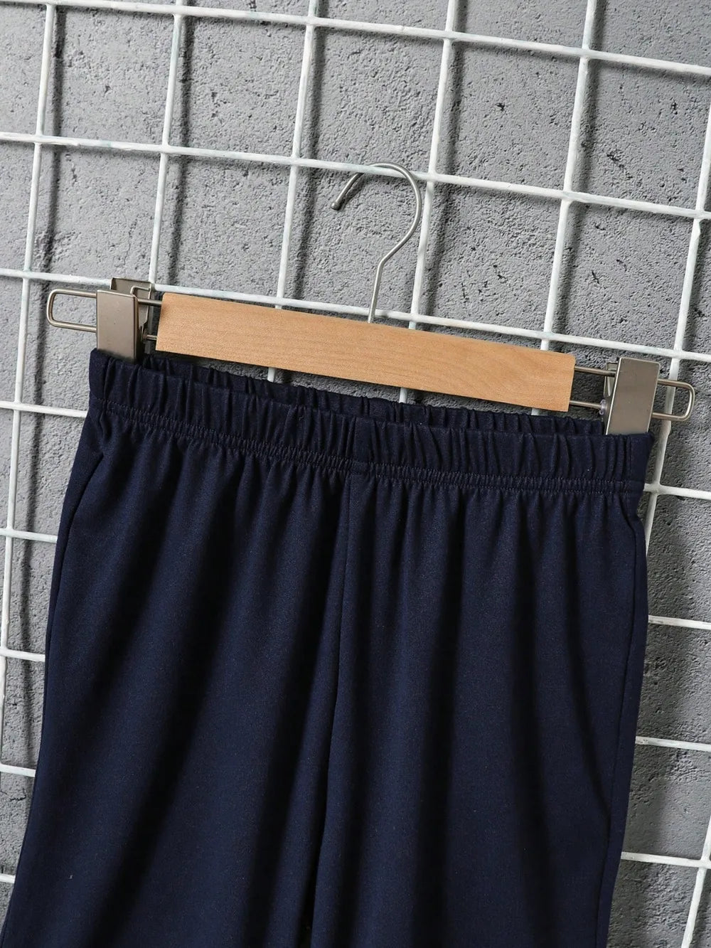 Navy sweatpants hanging on a wooden hanger against a gray grid backdrop.