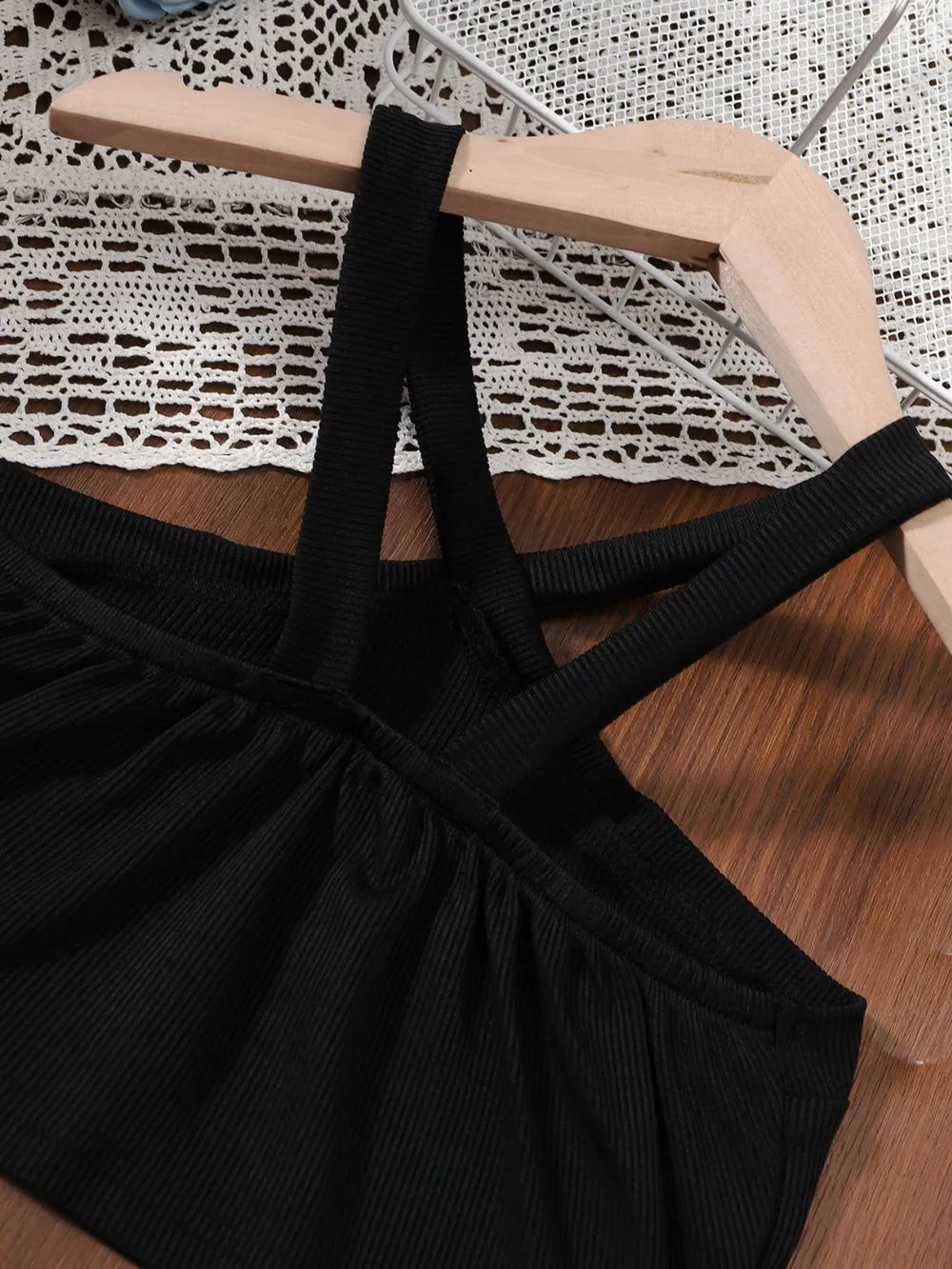 Black crisscross tank top hanging on a wooden hanger against a lace backdrop.
