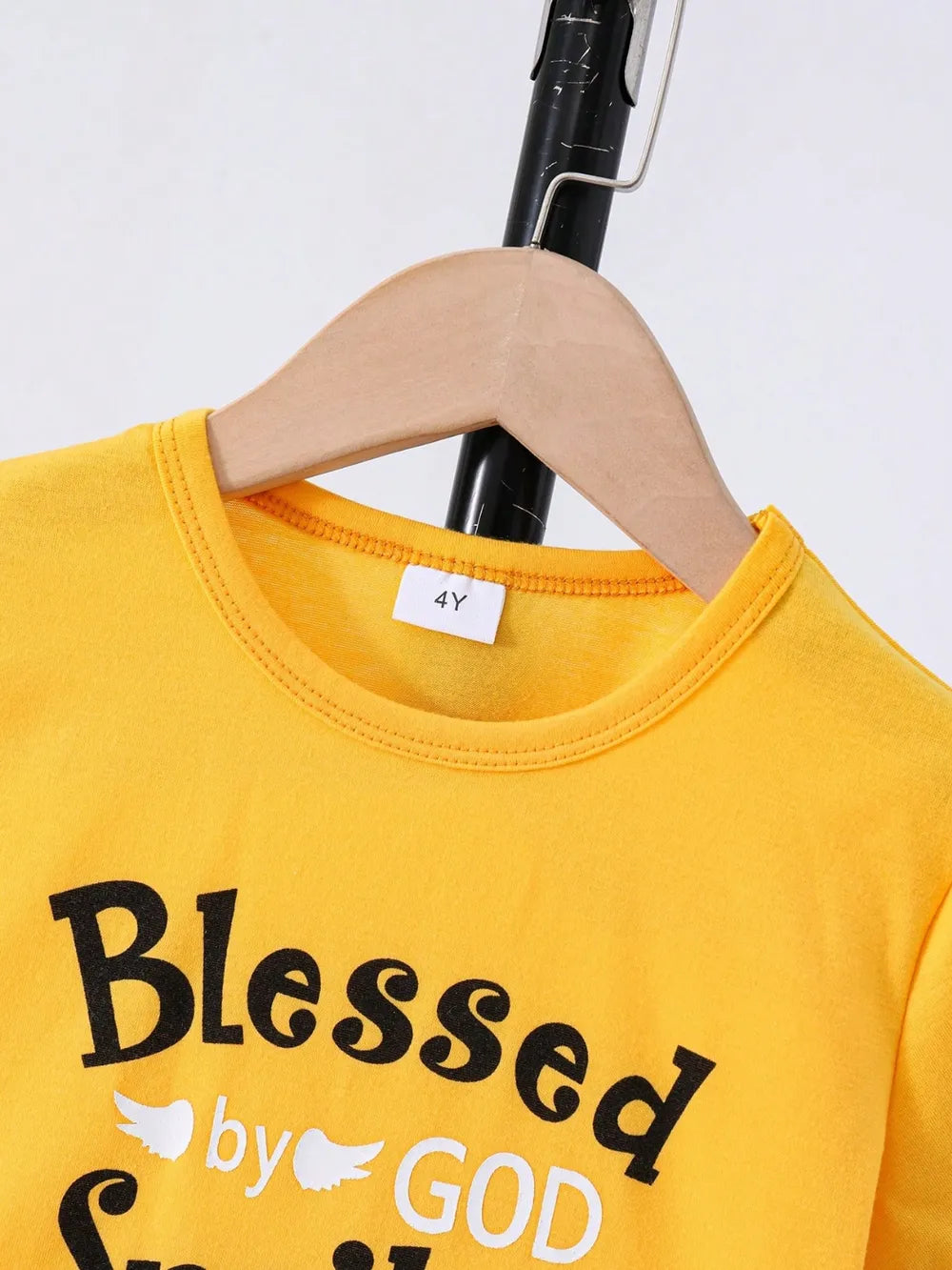 Yellow children's t-shirt on a wooden hanger, with "Blessed by God" printed in black and white.