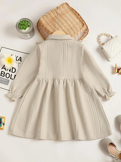 Light beige children's dress with ruffled collar and bow, displayed on a wooden hanger.